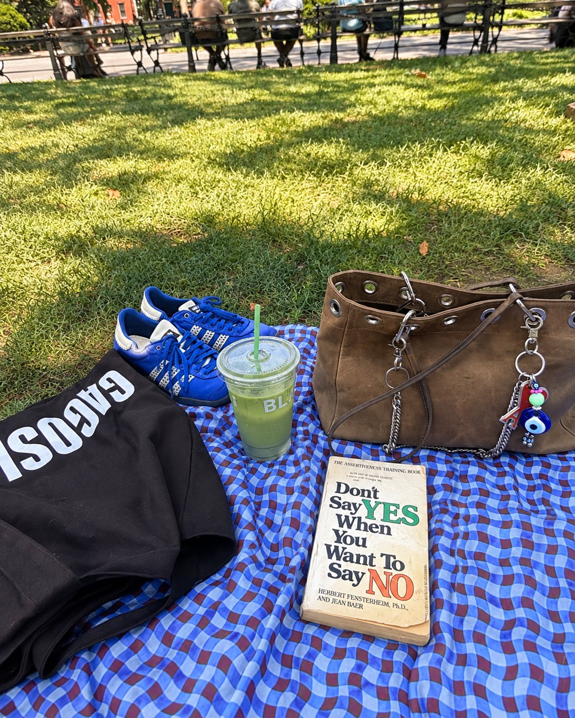 A picnic blanket in a park with a book, a green juice, a tote bag and blue sneakers