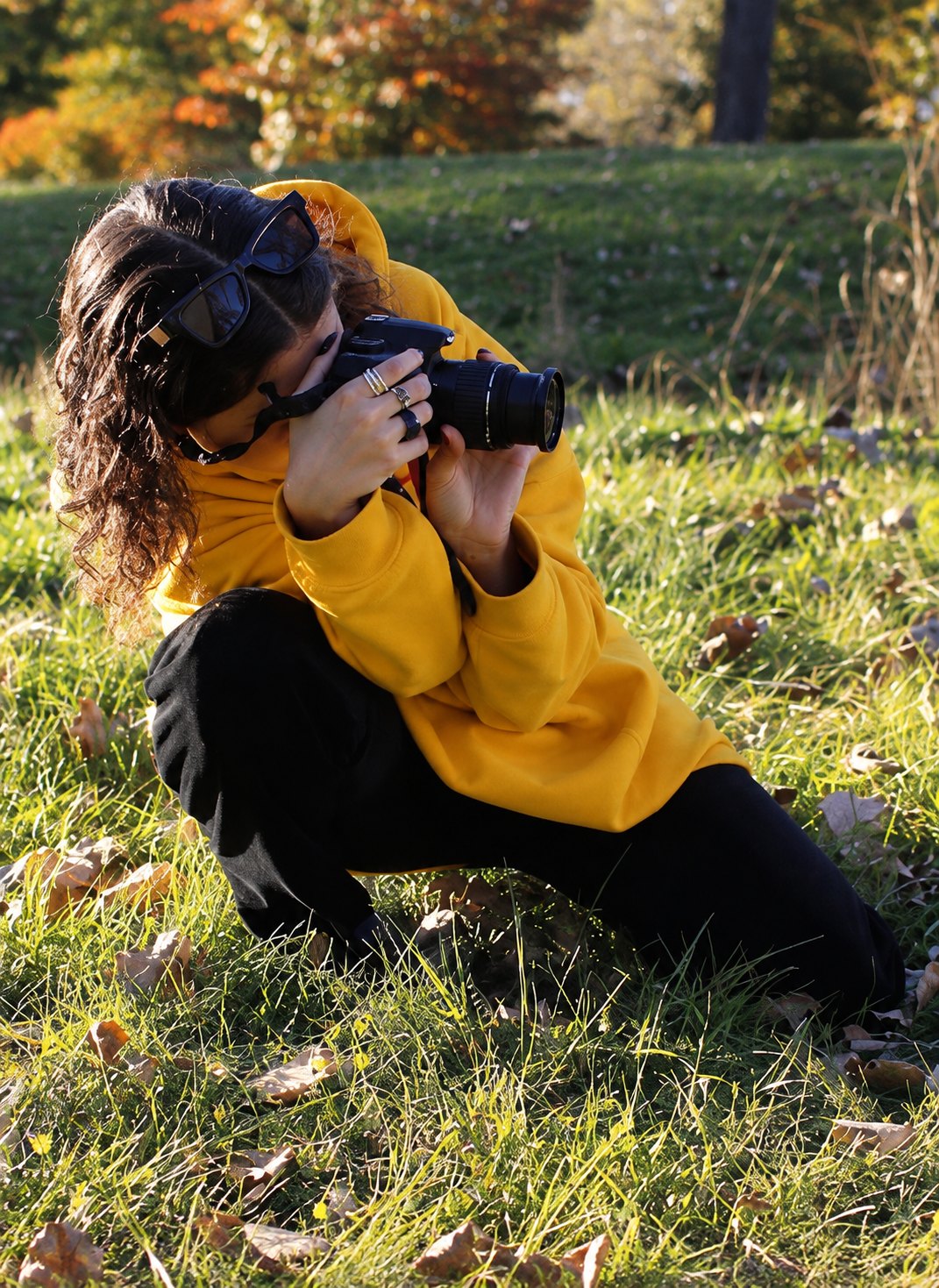 Aliya crouched in autumn grass in a yellow hoodie, shooting with a DSLR, sunglasses pushed up into her curls