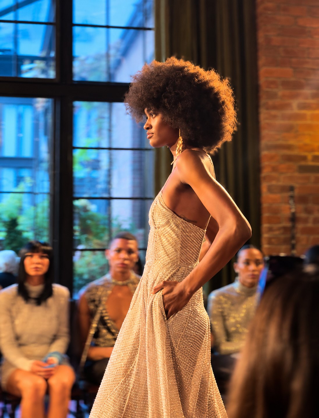 A model with a full afro walking the Brunello Cucinelli runway in a beaded silver gown, hand in pocket, seated guests behind her