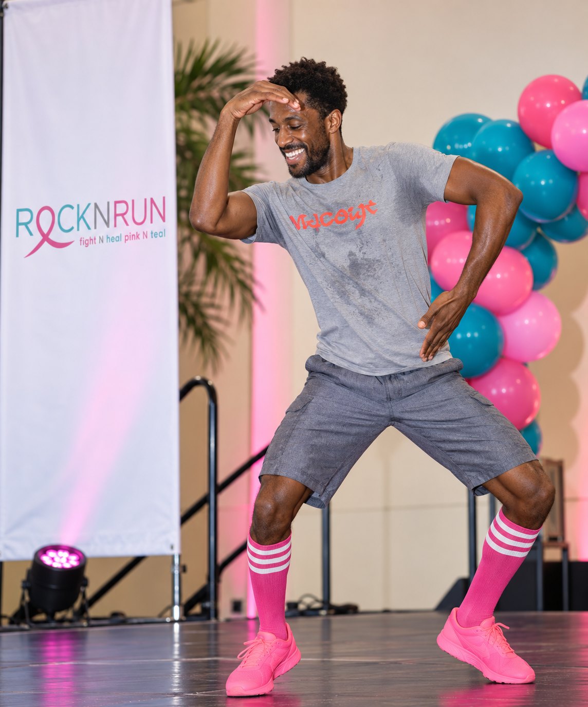 A dance instructor in a grey shirt and pink knee socks mid-move on stage beside a Rock N Run banner and pink and teal balloons