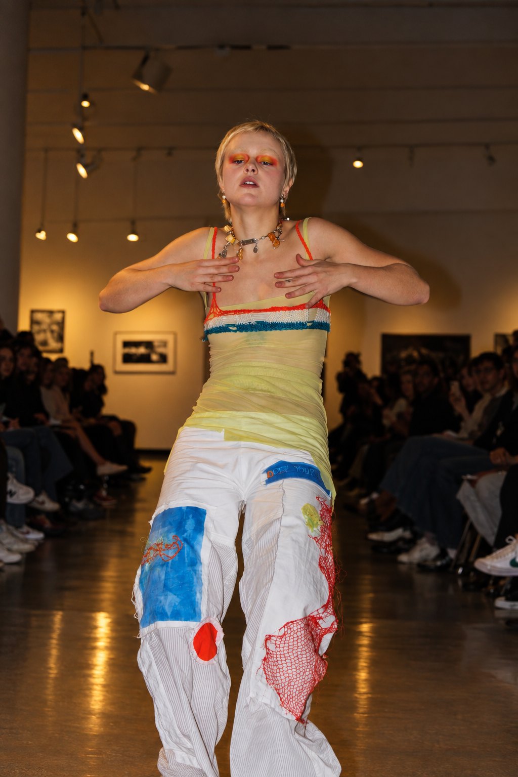 A short-haired model dancing down the runway in a yellow top and white patchwork trousers