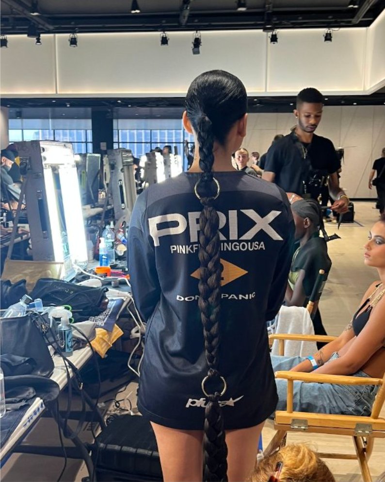 A model backstage in a black jersey with a waist-length braid, mirrors and stylists behind her