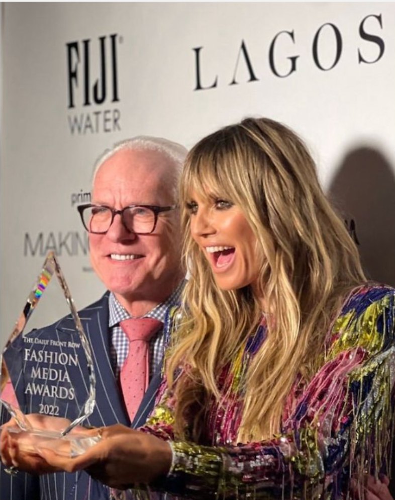 Two award recipients holding a Fashion Media Awards trophy in front of a sponsor step and repeat