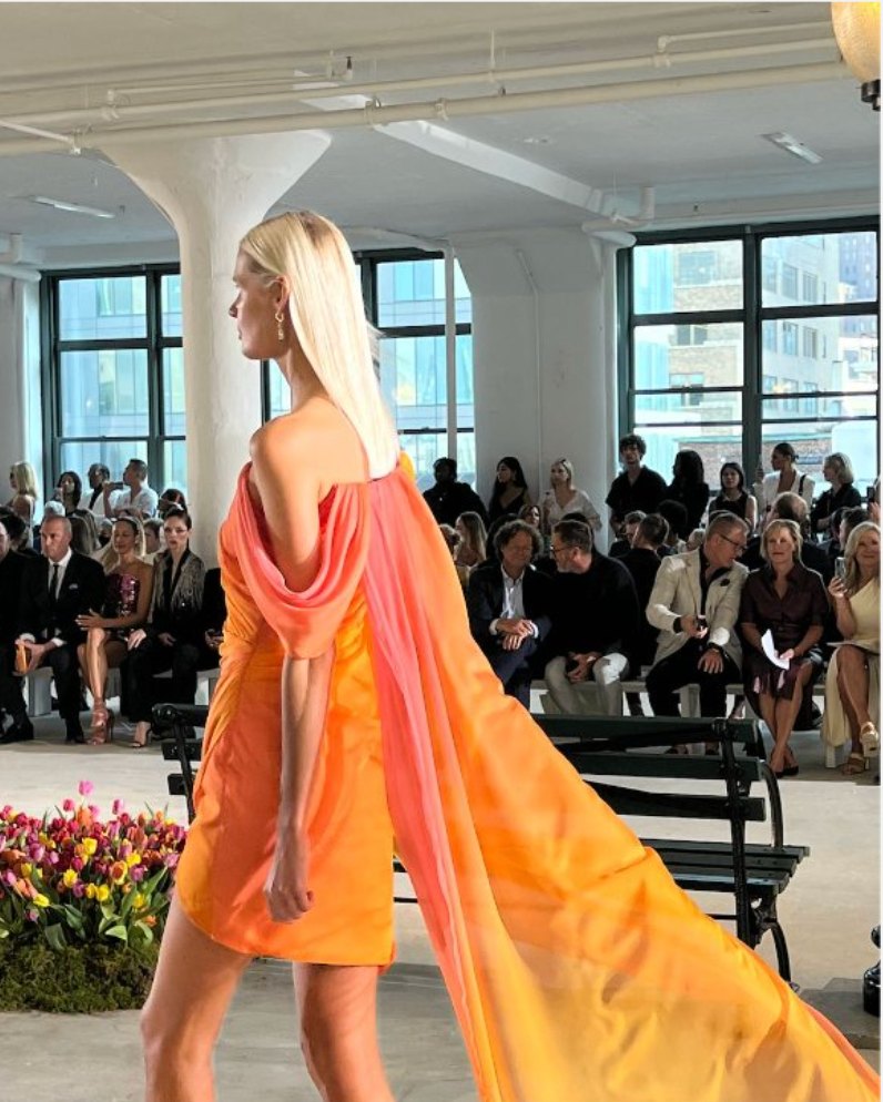 A model walking a NYFW runway in an orange draped dress with a long chiffon train