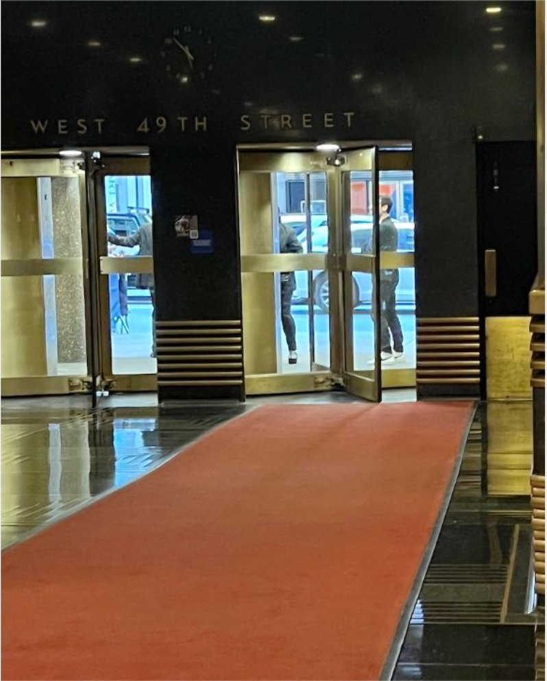An empty red carpet running to brass revolving doors under a West 49th Street sign