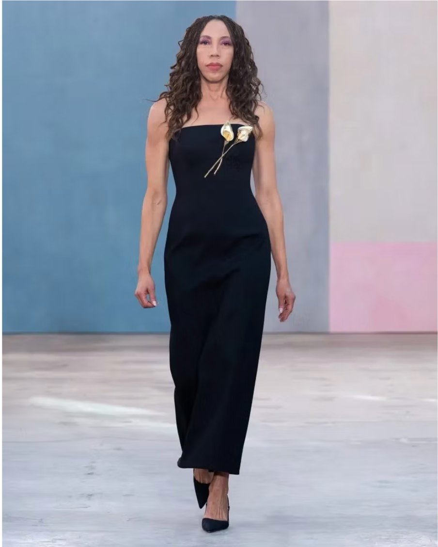 The painter Amy Sherald walking a runway in a black column dress with two gold calla lilies at the bodice