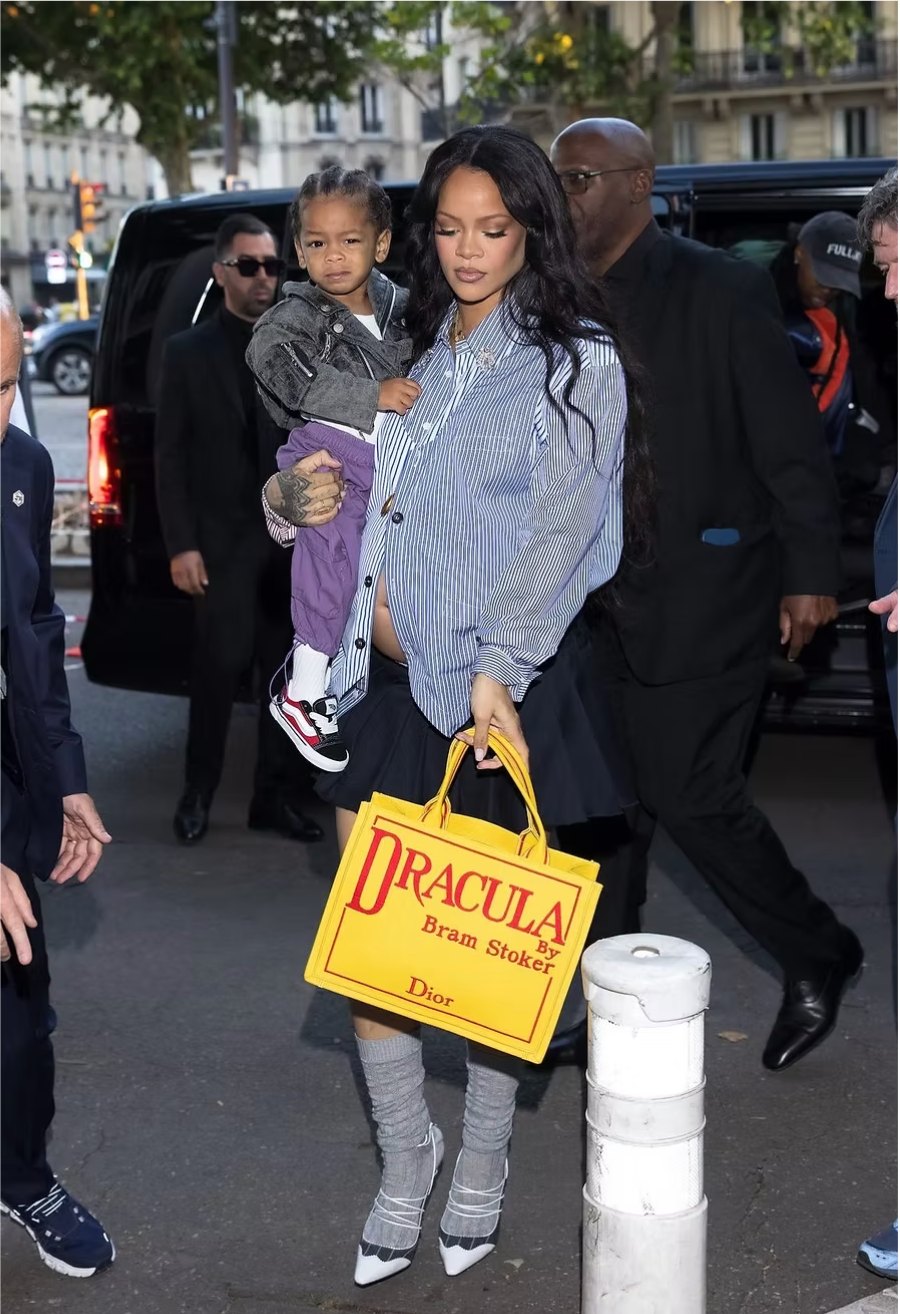 Rihanna carrying a yellow Dior tote printed as a Dracula book cover while holding her son on a Paris street
