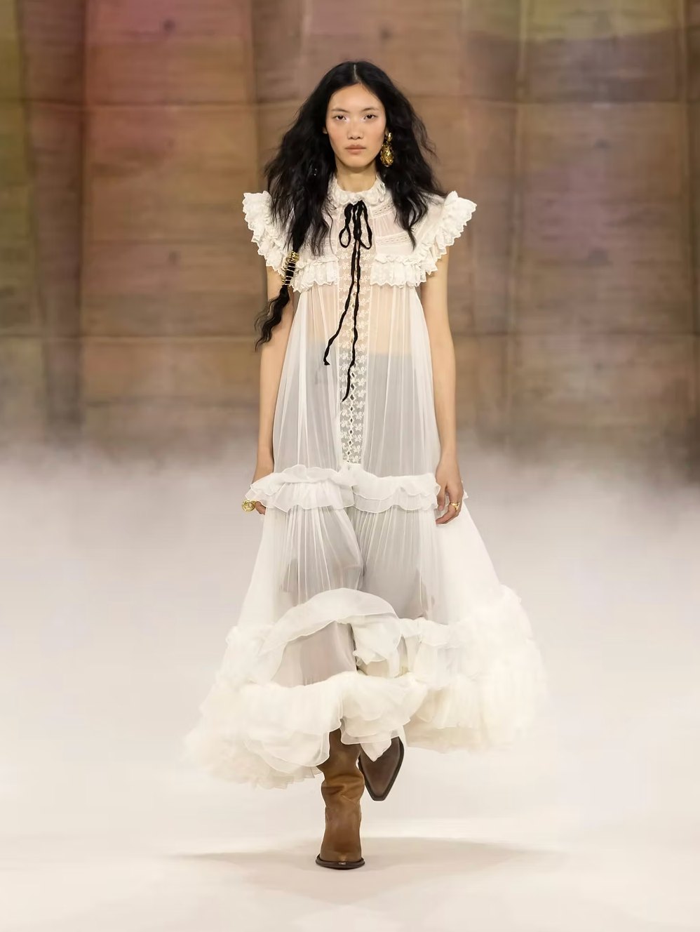A Chloe look of a sheer tiered white ruffled dress with a black ribbon tie, worn with tan cowboy boots