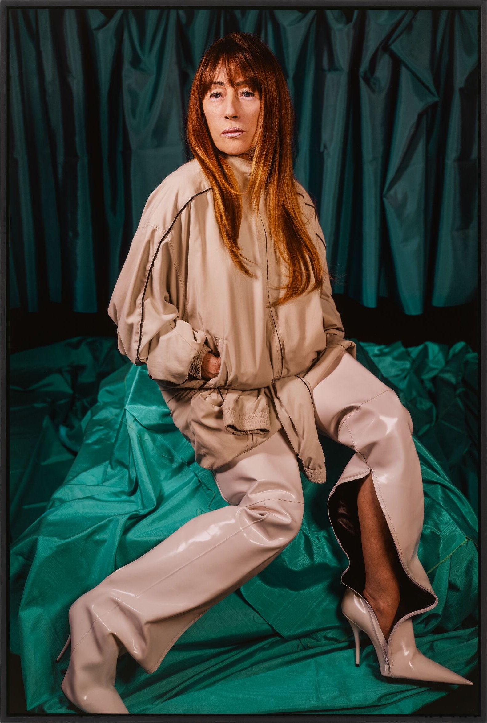A photograph of a red-haired woman in a beige bomber jacket and pale latex boots seated on green fabric before a dark green curtain