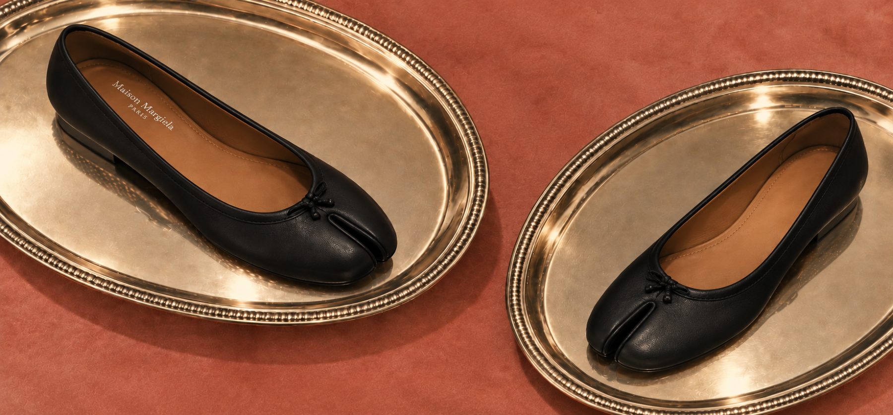 Two black split-toe ballet flats photographed on gold oval platters against a terracotta ground