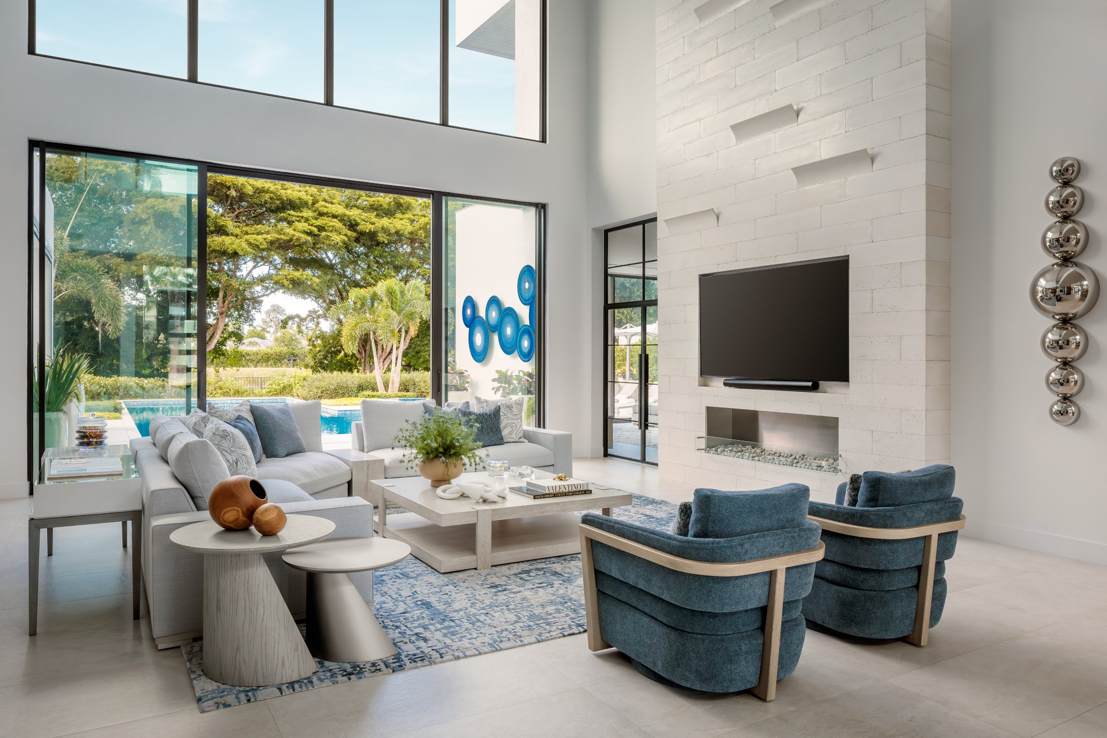 A double-height living room with blue lounge chairs, opening through tall glass doors to a private pool garden